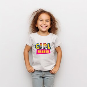Peut inclure: Un t-shirt blanc avec le mot "GIRL" en lettres bleues et jaunes, et le nom "JESSIE" en lettres roses. La chemise est port&eacute;e par une jeune fille aux cheveux bruns boucl&eacute;s et un jean bleu.