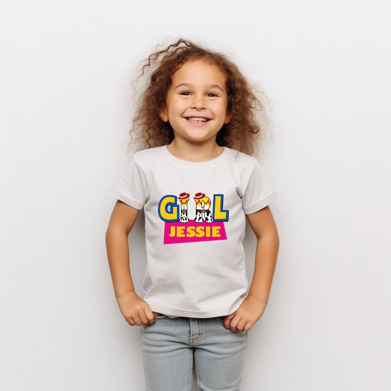Peut inclure: Un t-shirt blanc avec le mot "GIRL" en lettres bleues et jaunes, et le nom "JESSIE" en lettres roses. La chemise est port&eacute;e par une jeune fille aux cheveux bruns boucl&eacute;s et un jean bleu.