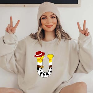 Peut inclure: Un sweat-shirt beige avec un motif de lettre "U" color&eacute;. Le "U" est orn&eacute; d'un chapeau de cow-boy, d'accents jaunes et d'un motif de vache. Le sweat-shirt est de couleur neutre et a des manches longues.