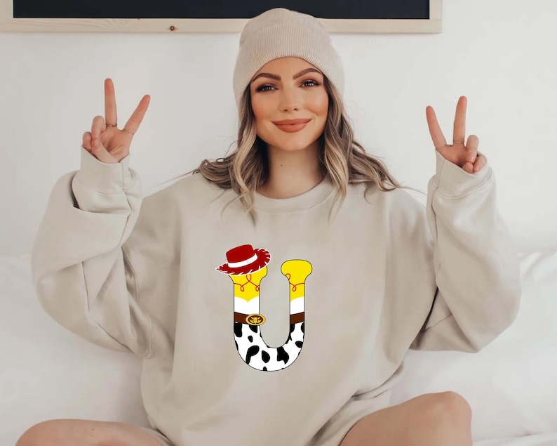 Peut inclure: Un sweat-shirt beige avec un motif de lettre "U" color&eacute;. Le "U" est orn&eacute; d'un chapeau de cow-boy, d'accents jaunes et d'un motif de vache. Le sweat-shirt est de couleur neutre et a des manches longues.