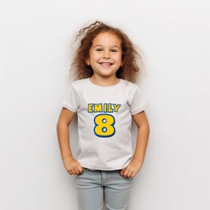 Peut inclure: Un t-shirt blanc avec le nom "EMILY" en lettres bleues et jaunes et le chiffre "8" dans un style similaire. La chemise est port&eacute;e par une jeune fille aux cheveux bruns boucl&eacute;s et un jean bleu.