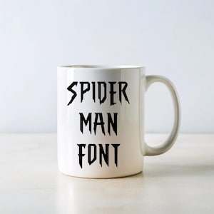 May include: White ceramic mug with the words "SPIDER MAN FONT" in black, stylized lettering. The mug has a curved handle and a simple, clean design. The text is arranged vertically on the mug.