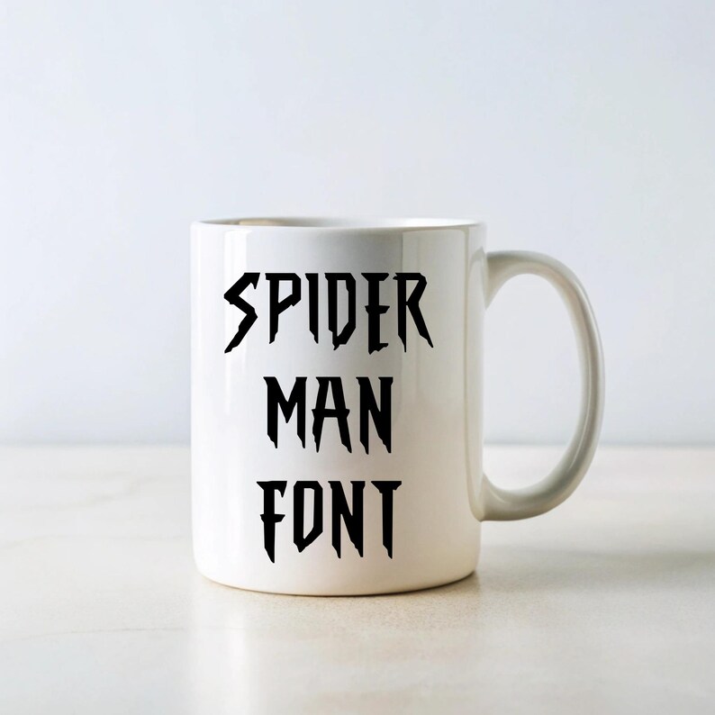 May include: White ceramic mug with the words "SPIDER MAN FONT" in black, stylized lettering. The mug has a curved handle and a simple, clean design. The text is arranged vertically on the mug.