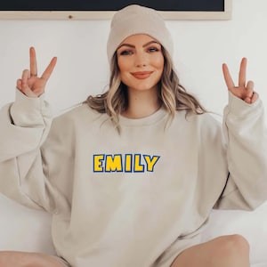 Peut inclure: Un sweat-shirt beige avec le nom "EMILY" en lettres bleues avec un contour jaune. La personne sur l'image porte un bonnet beige et fait le signe de la paix avec les deux mains.