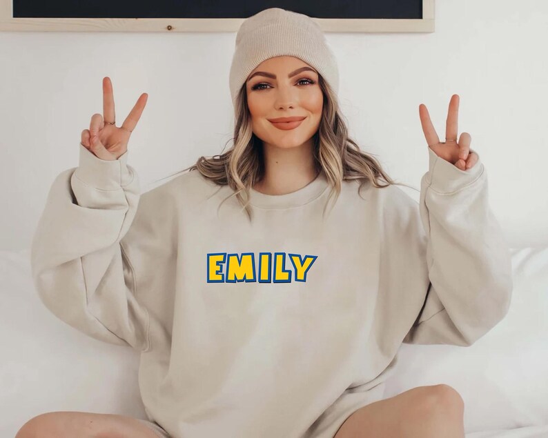 Peut inclure: Un sweat-shirt beige avec le nom "EMILY" en lettres bleues avec un contour jaune. La personne sur l'image porte un bonnet beige et fait le signe de la paix avec les deux mains.