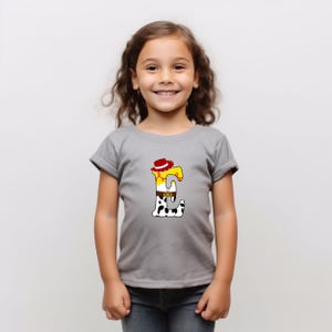 Peut inclure: Un t-shirt gris avec un motif color&eacute; de la lettre "E". Le motif comprend un chapeau de cowboy, une chemise jaune, une ceinture et un pantalon &agrave; imprim&eacute; vache. Le t-shirt est port&eacute; par une jeune fille.