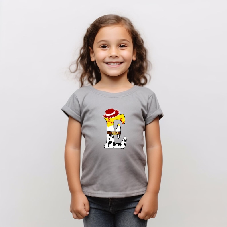 Peut inclure: Un t-shirt gris avec un motif color&eacute; de la lettre "E". Le motif comprend un chapeau de cowboy, une chemise jaune, une ceinture et un pantalon &agrave; imprim&eacute; vache. Le t-shirt est port&eacute; par une jeune fille.