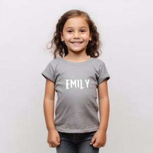 May include: A gray t-shirt with the name "EMILY" printed in white, worn by a young girl. The shirt has short sleeves and a crew neck. The girl is wearing blue jeans.