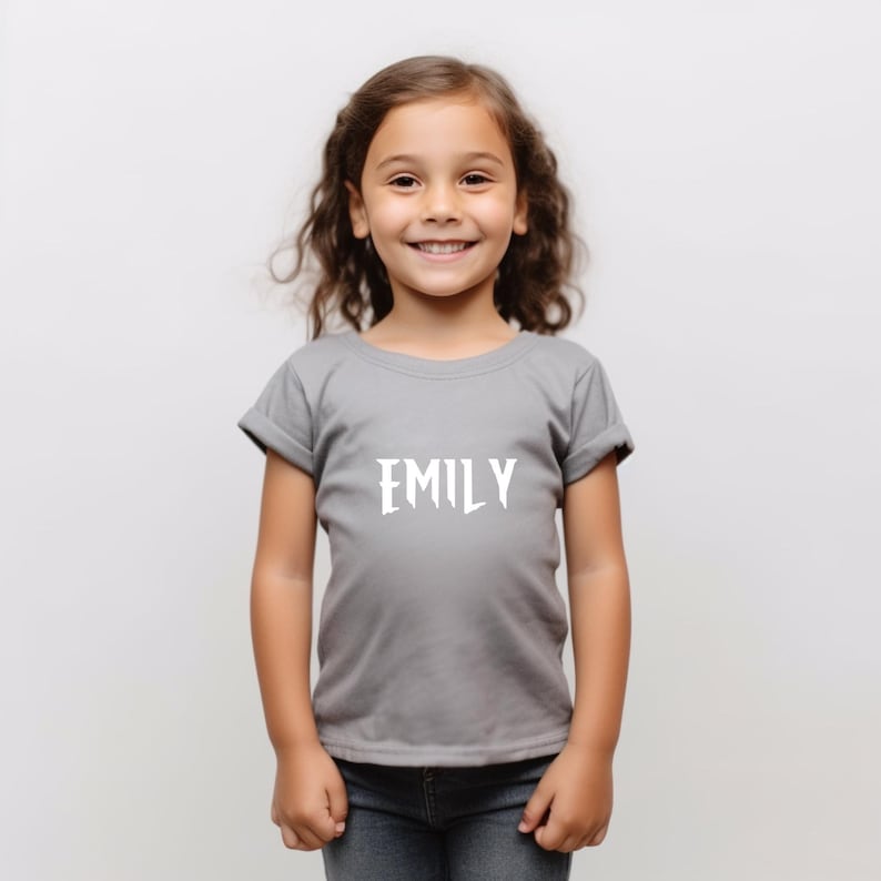 May include: A gray t-shirt with the name "EMILY" printed in white, worn by a young girl. The shirt has short sleeves and a crew neck. The girl is wearing blue jeans.