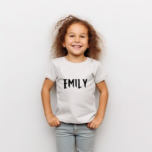 May include: A white t-shirt with the name "EMILY" printed in black, stylized font. The shirt is worn by a young child with curly brown hair and blue jeans.