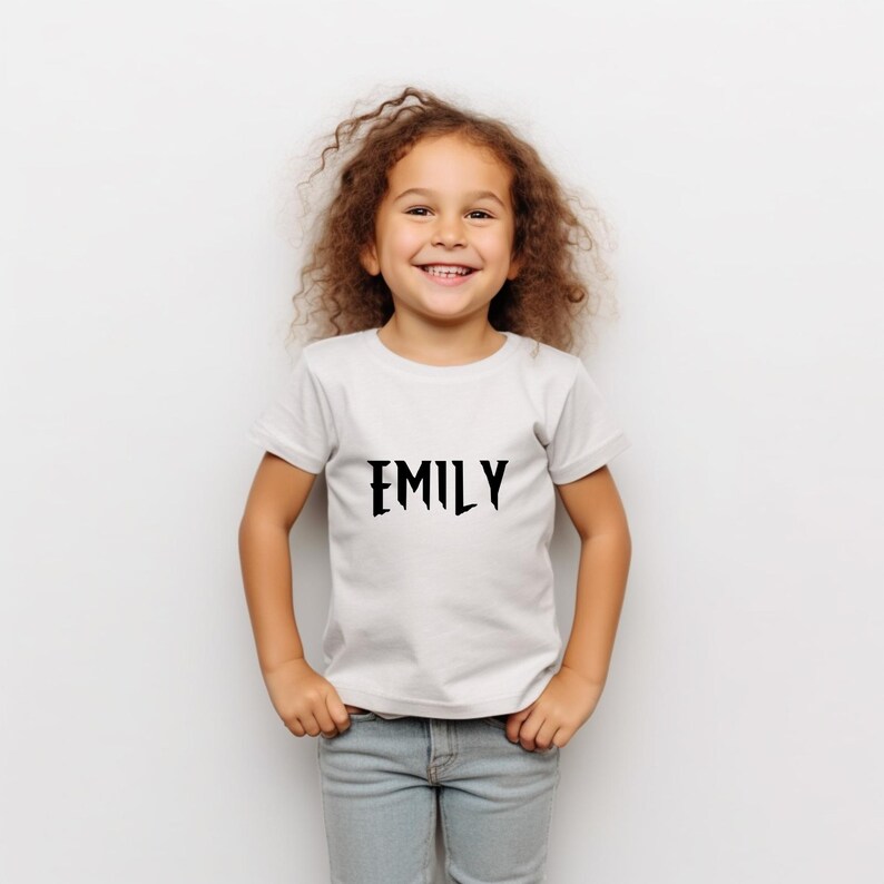 May include: A white t-shirt with the name "EMILY" printed in black, stylized font. The shirt is worn by a young child with curly brown hair and blue jeans.