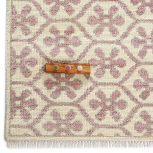 Op de afbeelding: Een gedeelte van een crèmekleurig tapijt met een herhalend bloemenpatroon in lichtpaars. Een houten kapstok met drie gekleurde pinnen is op het tapijt geplaatst. Het tapijt heeft een gefranjerde rand.