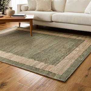 May include: A rectangular area rug with a green center and a beige border sits on a hardwood floor. The rug is in front of a white sofa with beige pillows. A wooden coffee table and plants are also in the room.