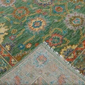 Op de afbeelding: Een groen vloerkleed met een ingewikkeld bloemenpatroon in tinten oranje, blauw en geel. Het vloerkleed heeft een gedetailleerde rand en een gefranjerde rand. Het vloerkleed is opgevouwen en toont de onderkant.