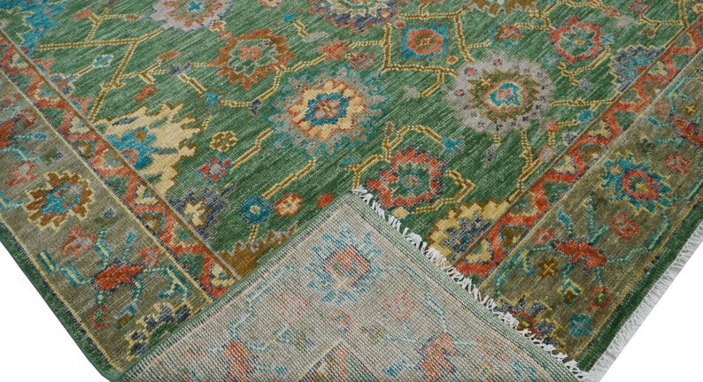 Op de afbeelding: Een groen vloerkleed met een ingewikkeld bloemenpatroon in tinten oranje, blauw en geel. Het vloerkleed heeft een gedetailleerde rand en een gefranjerde rand. Het vloerkleed is opgevouwen en toont de onderkant.