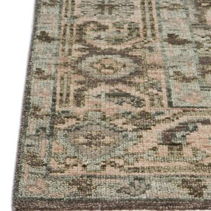 May include: Close-up of a rectangular rug with a detailed, faded pattern. The rug features a combination of muted colors, including dusty rose, sage green, and brown. The edges are finished with a fringe.