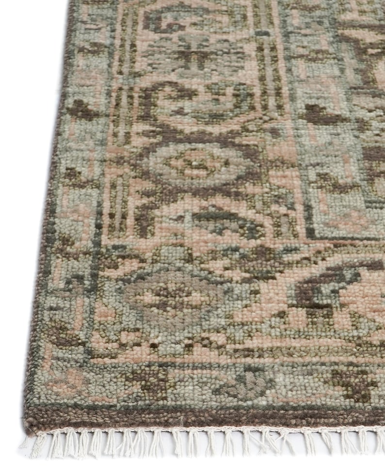 May include: Close-up of a rectangular rug with a detailed, faded pattern. The rug features a combination of muted colors, including dusty rose, sage green, and brown. The edges are finished with a fringe.