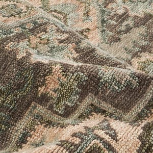 May include: Close-up of a rolled-up area rug with a detailed, textured pattern. The rug features a blend of colors, including shades of brown, green, and beige. The design appears to be a traditional, possibly oriental, motif.