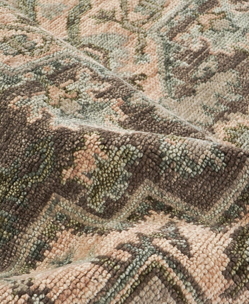 May include: Close-up of a rolled-up area rug with a detailed, textured pattern. The rug features a blend of colors, including shades of brown, green, and beige. The design appears to be a traditional, possibly oriental, motif.
