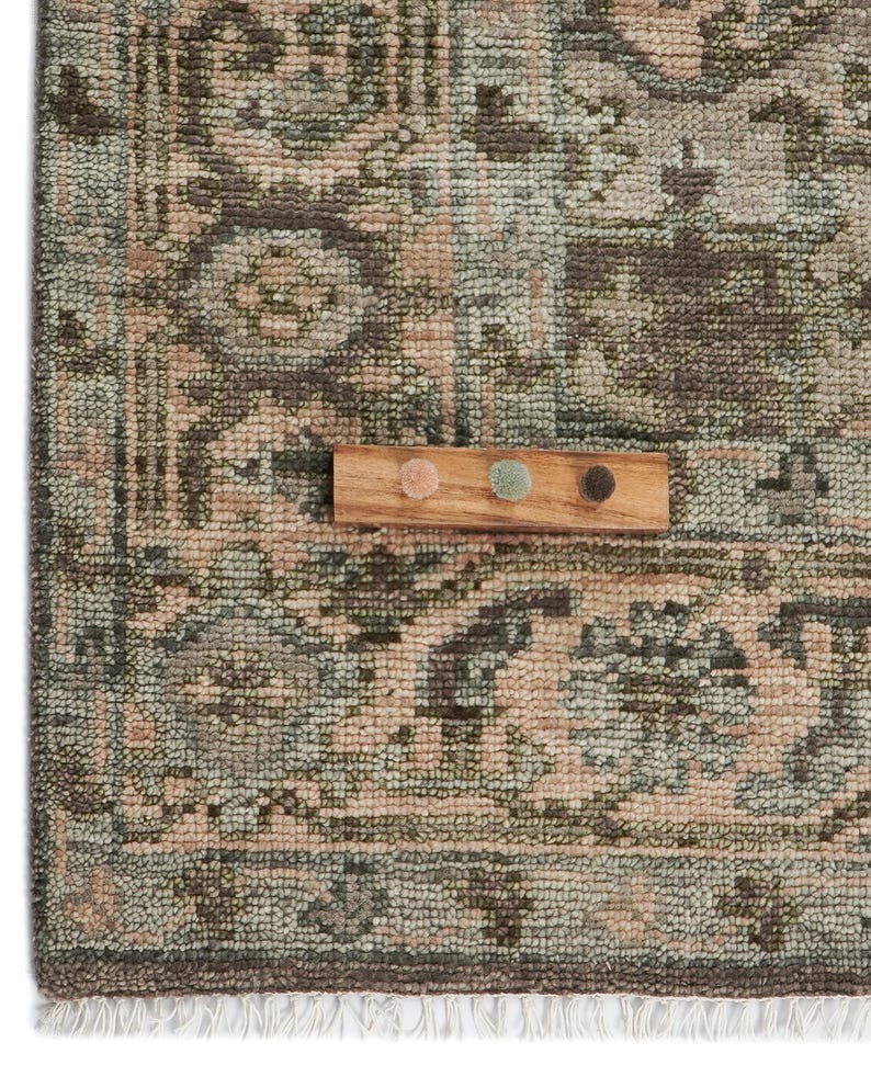 May include: Close-up of a patterned rug with a distressed, vintage look. The rug features a mix of muted colors, including green, brown, and pink. A small wooden swatch board with three color samples sits on top of the rug, showcasing the color palette.