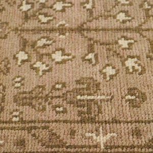 以下が含まれることがあります： くすんだピンクのベースカラーに、茶色とクリーム色の花柄や幾何学模様が繰り返される織りラグ。厚みのある結び目のある質感で、ラグの下端に沿って明確な縁取り部分が見えるデザインとなっている。