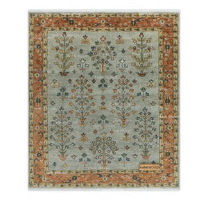 Puede incluir: Una alfombra rectangular con un campo gris claro y un patrón repetitivo de árboles florales estilizados en tonos naranja, azul y verde. La alfombra tiene un borde ancho de color naranja con motivos florales y una pequeña etiqueta rectangular HANKWOVEN.
