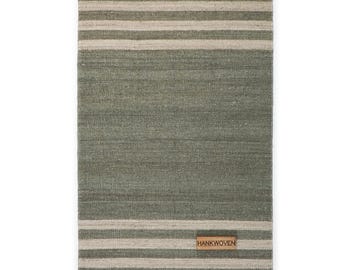May include: A rectangular, woven rug in shades of green and beige. The rug features horizontal stripes of beige against a green background. A small, rectangular leather tag with the text "HANKWOVEN" is attached to the rug.