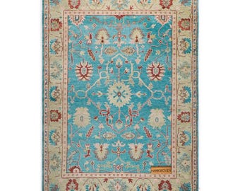 May include: A rectangular turquoise rug with an intricate floral pattern. The rug features a central turquoise field with cream and red floral motifs. A cream border with red and blue floral designs frames the central field. The rug has the text "HANKWOVEN".