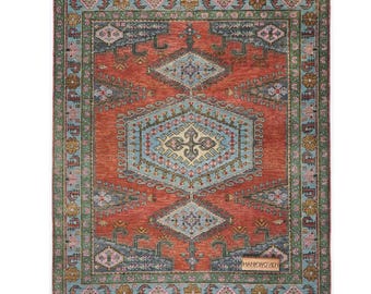 May include: Rectangular rug with a central geometric design in shades of red, blue, and cream. The rug features a detailed border in green and blue, with a label that reads "HANKWOVEN".