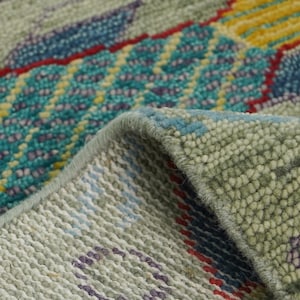 Op de afbeelding: Close-up van een deel van een tapijt met een gedetailleerd, gestructureerd patroon. Het tapijt heeft een mix van kleuren, waaronder groentinten, blauw, geel en rood. Het ontwerp bevat geometrische en abstracte elementen, met een zichtbare gevouwen rand.