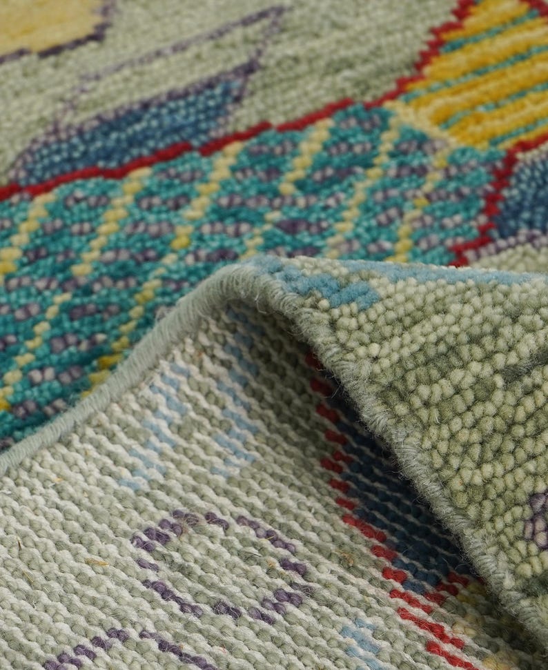 Op de afbeelding: Close-up van een deel van een tapijt met een gedetailleerd, gestructureerd patroon. Het tapijt heeft een mix van kleuren, waaronder groentinten, blauw, geel en rood. Het ontwerp bevat geometrische en abstracte elementen, met een zichtbare gevouwen rand.