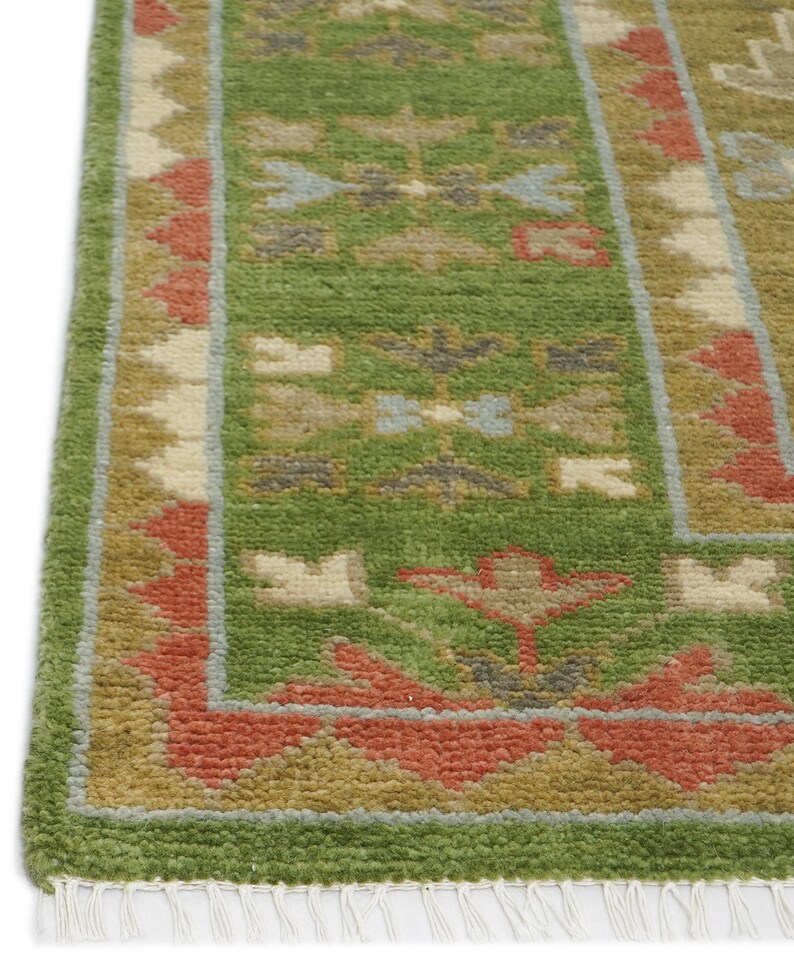 以下が含まれることがあります： 詳細な模様の緑色のラグのクローズアップ。ラグは、ベージュ、赤、青の色合いの幾何学模様の緑色の中心フィールドを備えています。ボーダーは緑、ベージュ、赤の混合で、端にフリンジが付いています。