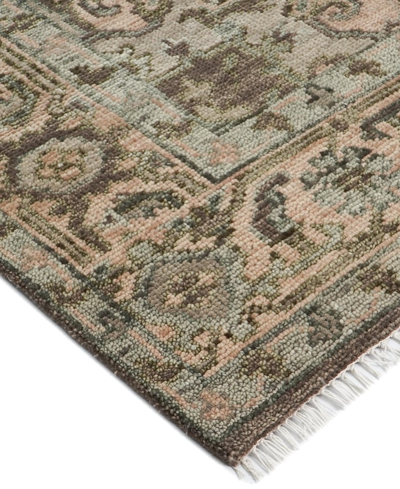 May include: Close-up of a section of a patterned rug. The rug features a traditional design with a mix of colors, including muted pink, green, and brown tones. The edges are finished with a dark brown border and white fringe.