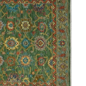 Op de afbeelding: Een detail van een groene vloerkleed met een ingewikkeld bloemenpatroon. Het kleed heeft een groen middenveld met een herhalend ontwerp in blauw, rood, geel en bruin. Een donkerdere groene rand omlijst het ontwerp, met franjes aan de rechterkant.