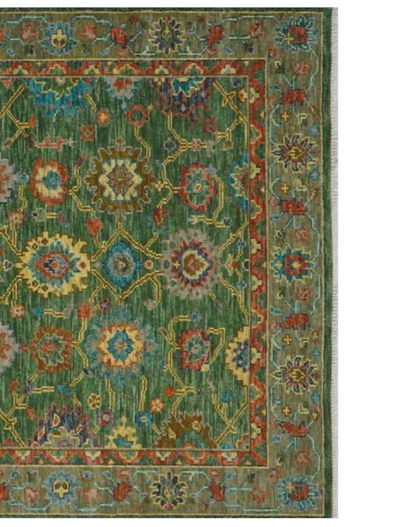 Op de afbeelding: Een detail van een groene vloerkleed met een ingewikkeld bloemenpatroon. Het kleed heeft een groen middenveld met een herhalend ontwerp in blauw, rood, geel en bruin. Een donkerdere groene rand omlijst het ontwerp, met franjes aan de rechterkant.
