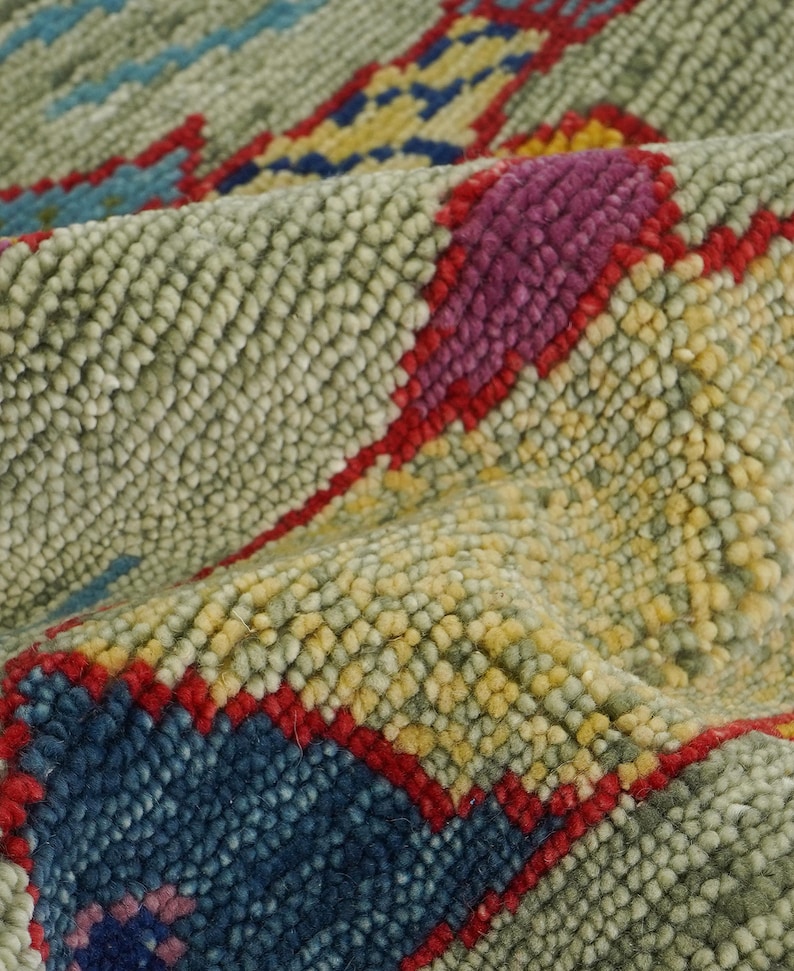 Op de afbeelding: Close-up van een getextureerd tapijt met een herhalend patroon. Het tapijt heeft een lichtgroene achtergrond met accenten van rood, blauw, geel en paars. Het ontwerp heeft een geometrische, tribale esthetiek.