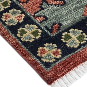 Puede incluir: Primer plano de una secci&oacute;n de alfombra con un patr&oacute;n floral detallado. La alfombra presenta un borde azul marino con motivos florales crema, verdes y rojos. El dise&ntilde;o central tiene elementos rojos, naranjas y verdes sobre un fondo verde azulado apagado. Flecos blancos visibles.