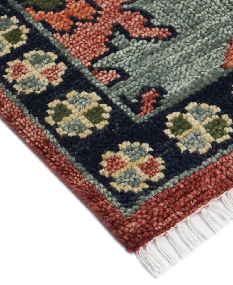 Puede incluir: Primer plano de una secci&oacute;n de alfombra con un patr&oacute;n floral detallado. La alfombra presenta un borde azul marino con motivos florales crema, verdes y rojos. El dise&ntilde;o central tiene elementos rojos, naranjas y verdes sobre un fondo verde azulado apagado. Flecos blancos visibles.