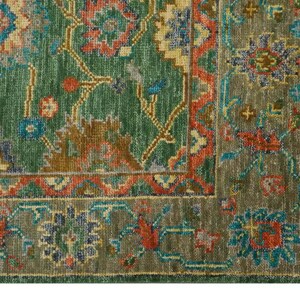 Op de afbeelding: Detail van een groen en roestkleurig tapijt met een ingewikkeld bloemenpatroon. Het tapijt heeft een verweerde uitstraling, met een franje langs de rand. Het patroon bevat tinten groen, roest, blauw en goud.