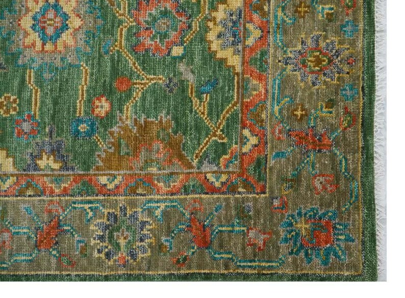 Op de afbeelding: Detail van een groen en roestkleurig tapijt met een ingewikkeld bloemenpatroon. Het tapijt heeft een verweerde uitstraling, met een franje langs de rand. Het patroon bevat tinten groen, roest, blauw en goud.