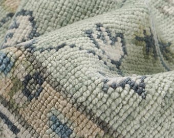 May include: Close-up of a woven rug with a detailed pattern. The rug features a mix of light green, blue, and beige colors. The texture is dense and plush, with visible knots and a slightly distressed appearance, suggesting a vintage style.