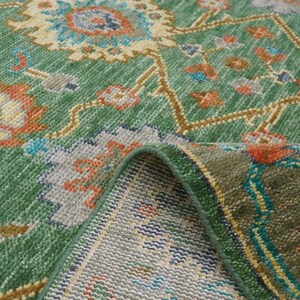 Op de afbeelding: Een groen vloerkleed met een gedetailleerd bloemenpatroon. Het vloerkleed heeft een centraal ontwerp met blauwe, oranje en gele bloemelementen op een groene achtergrond. De rand van het vloerkleed is omgevouwen en toont de onderkant.