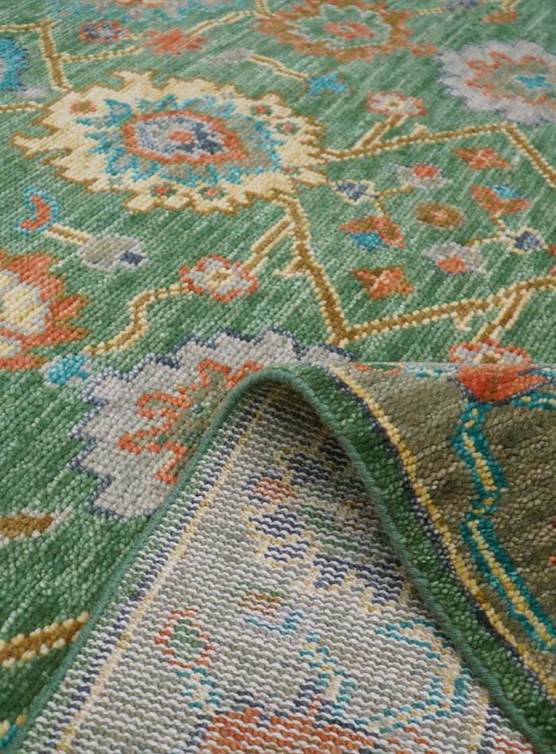 Op de afbeelding: Een groen vloerkleed met een gedetailleerd bloemenpatroon. Het vloerkleed heeft een centraal ontwerp met blauwe, oranje en gele bloemelementen op een groene achtergrond. De rand van het vloerkleed is omgevouwen en toont de onderkant.