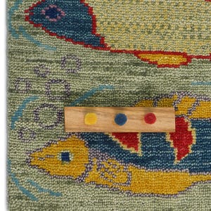Op de afbeelding: Een close-up van een kleurrijk tapijt met twee visontwerpen. De vissen zijn voornamelijk geel en blauw, met rode accenten, tegen een lichtgroene achtergrond. Een houten staal met gele, blauwe en rode stippen is op het tapijt geplaatst.
