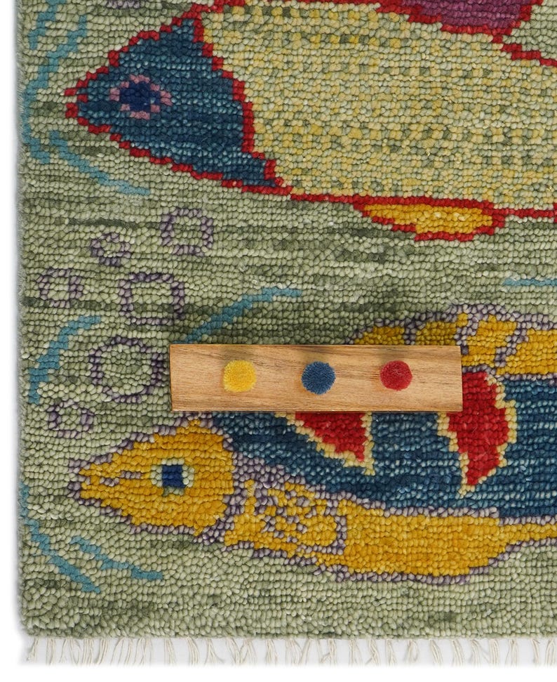Op de afbeelding: Een close-up van een kleurrijk tapijt met twee visontwerpen. De vissen zijn voornamelijk geel en blauw, met rode accenten, tegen een lichtgroene achtergrond. Een houten staal met gele, blauwe en rode stippen is op het tapijt geplaatst.