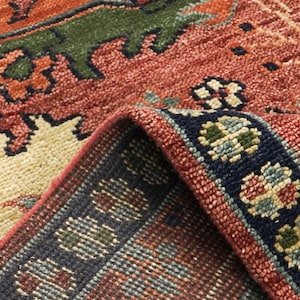 Puede incluir: Primer plano de una alfombra detallada y doblada con un dise&ntilde;o rico y texturizado. La alfombra presenta un tono rojo-naranja dominante con acentos de verde, azul marino y crema. El borde muestra un patr&oacute;n floral repetitivo. Los intrincados patrones y la paleta de colores de la alfombra crean una est&eacute;tica c&aacute;lida y acogedora.