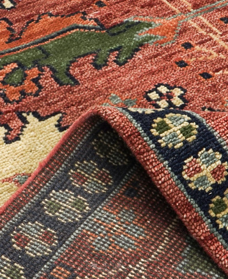 Puede incluir: Primer plano de una alfombra detallada y doblada con un dise&ntilde;o rico y texturizado. La alfombra presenta un tono rojo-naranja dominante con acentos de verde, azul marino y crema. El borde muestra un patr&oacute;n floral repetitivo. Los intrincados patrones y la paleta de colores de la alfombra crean una est&eacute;tica c&aacute;lida y acogedora.