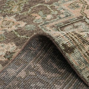 May include: Close-up of a rolled-up area rug with a detailed, faded floral pattern. The rug features a palette of muted colors, including brown, beige, and green. The underside of the rug is visible, showing a darker, textured backing.