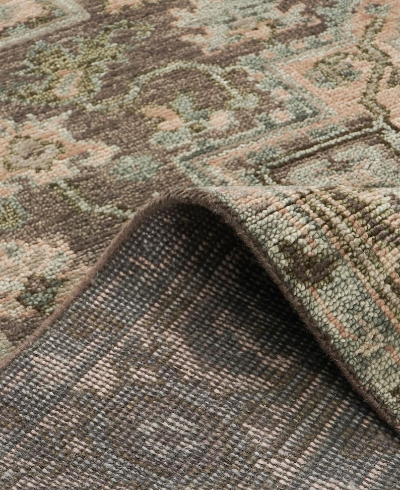 May include: Close-up of a rolled-up area rug with a detailed, faded floral pattern. The rug features a palette of muted colors, including brown, beige, and green. The underside of the rug is visible, showing a darker, textured backing.