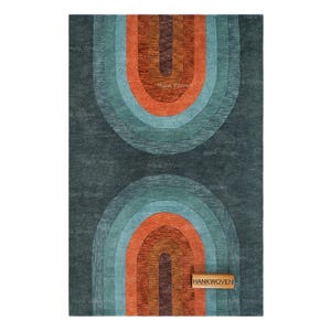 May include: A rectangular rug featuring a dark charcoal background with two symmetrical, stacked arch patterns in shades of burnt orange, teal, and deep brown. A small rectangular wooden tag with the text HANKWOVEN is attached to the lower right.